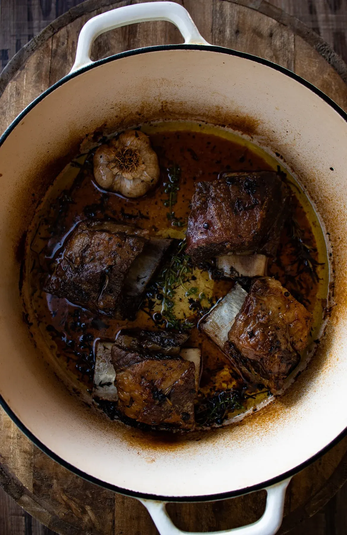 Bone-in beef short ribs braising in a cream-colored enamel Dutch oven viewed