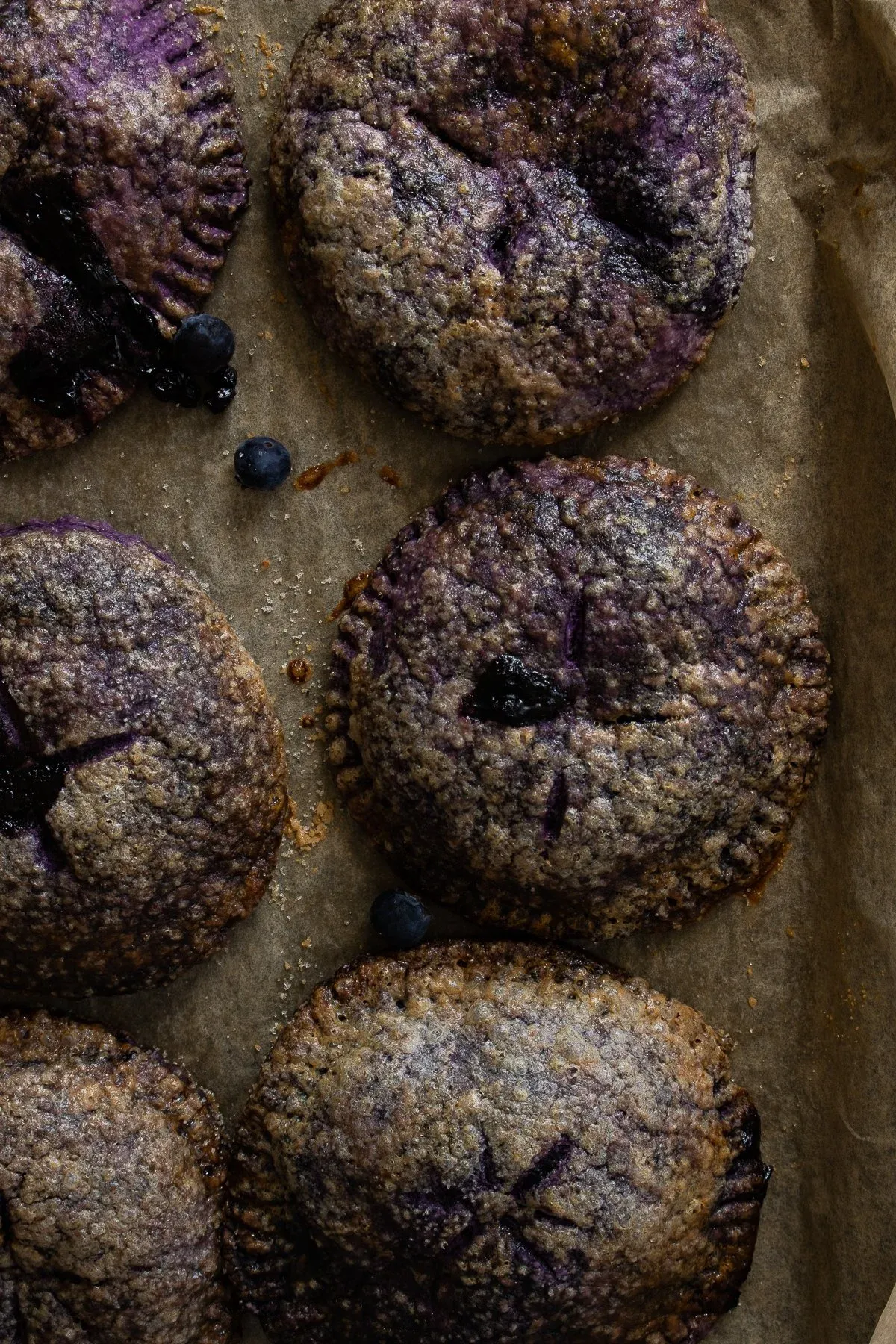Close overhead view of four Ube and Blueberry Mini Pies on parchment paper in moody dark directional lighting