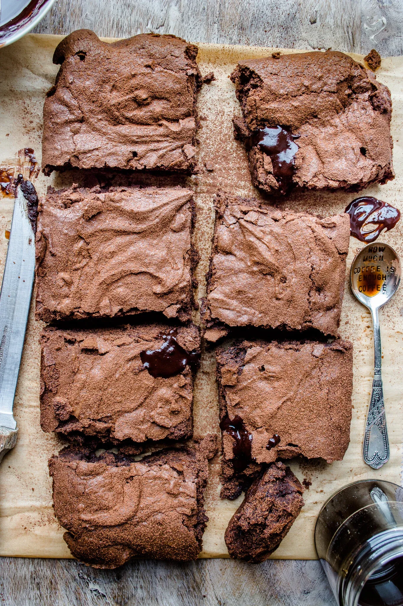Wider overhead shot of six fudgy gluten free espresso brownies on parchment paper atop a rustic wood surface