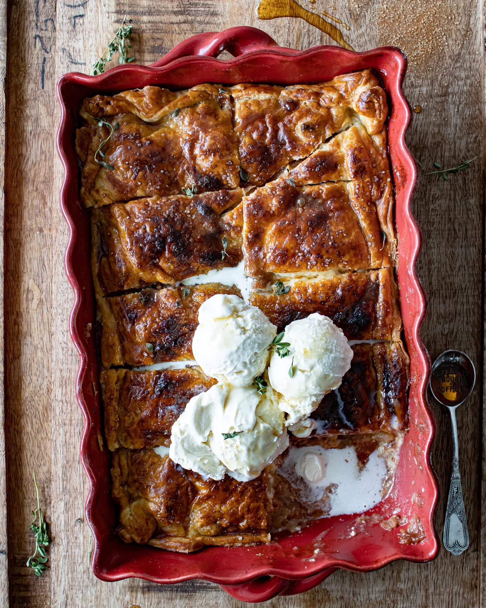 Apple and Bourbon Pandowdy in a red scalloped baking dish topped with three large scoops of melting vanilla ice cream nestled into the caramelized scored puff pastry