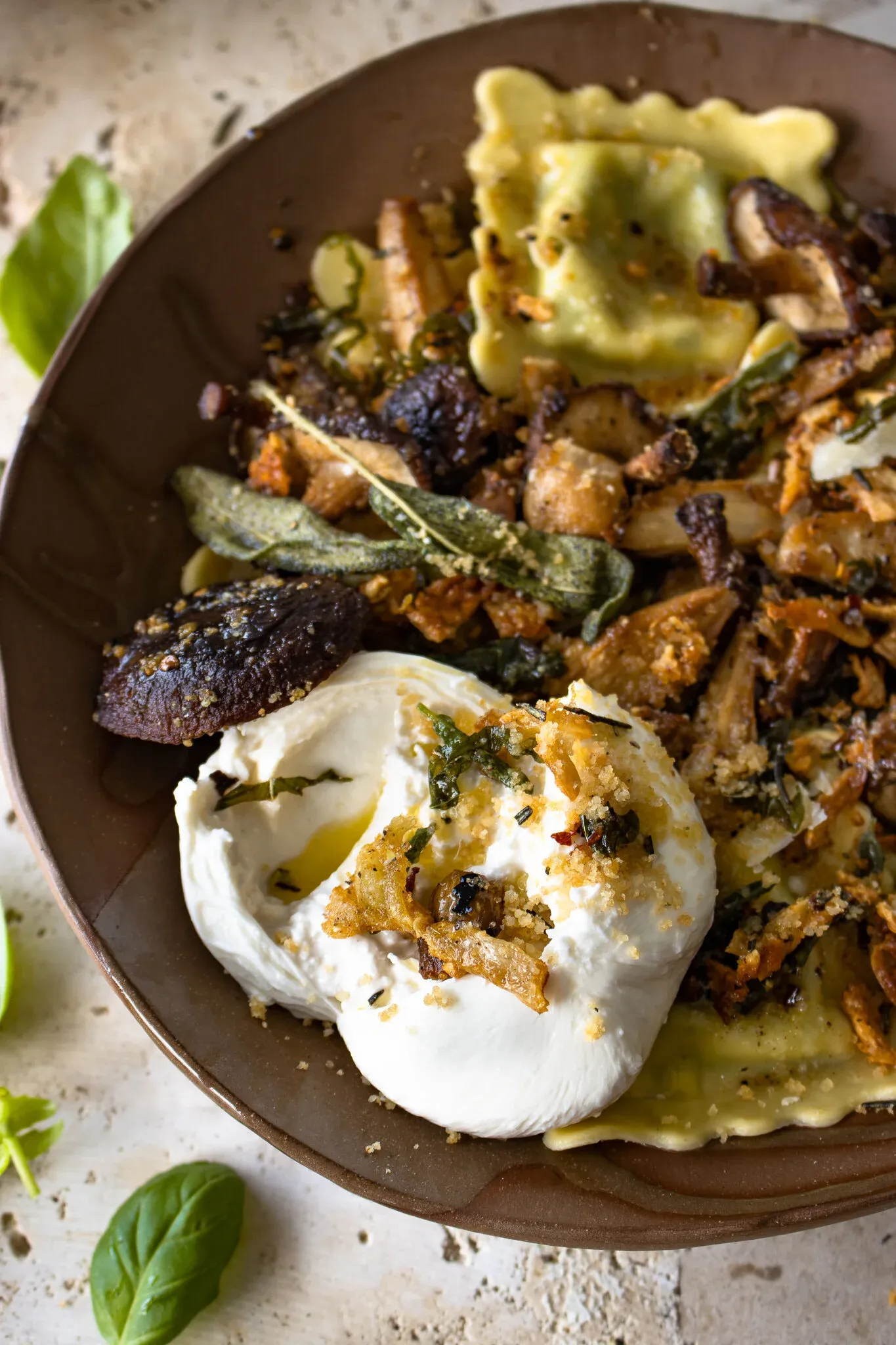 Close-up angled view of ravioli with roasted mushrooms in a brown bowl, showing the egg-yellow ravioli squares