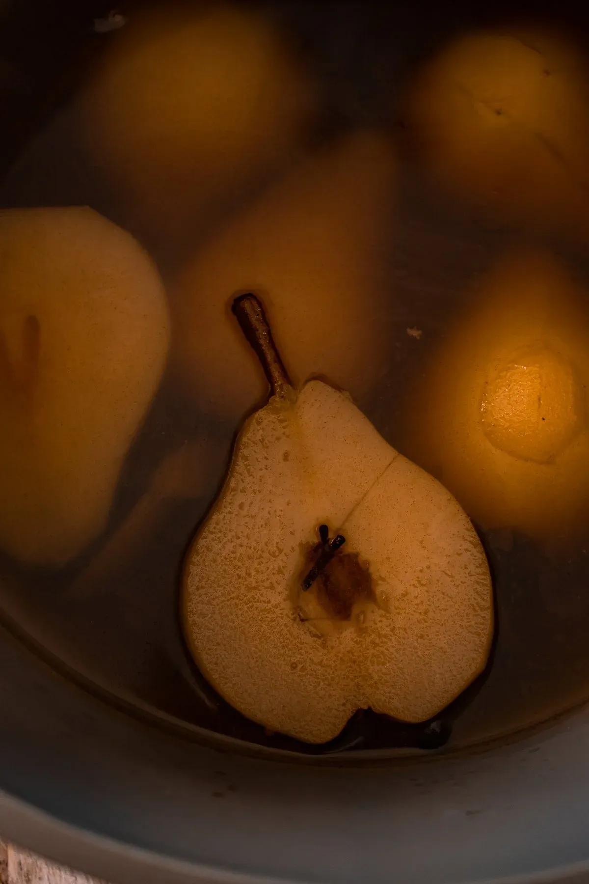 Close-up low-light shot of whole and halved pears submerged in amber poaching liquid inside a dark pot