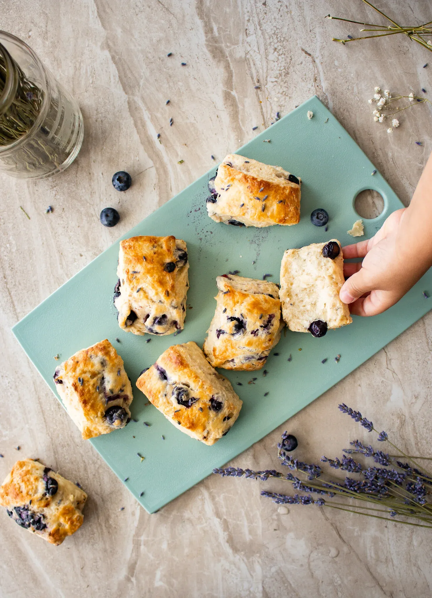 A hand reaches from the right to pick up one of the golden Blueberry Lavender Buttermilk Biscuits from the mint-green cutting board