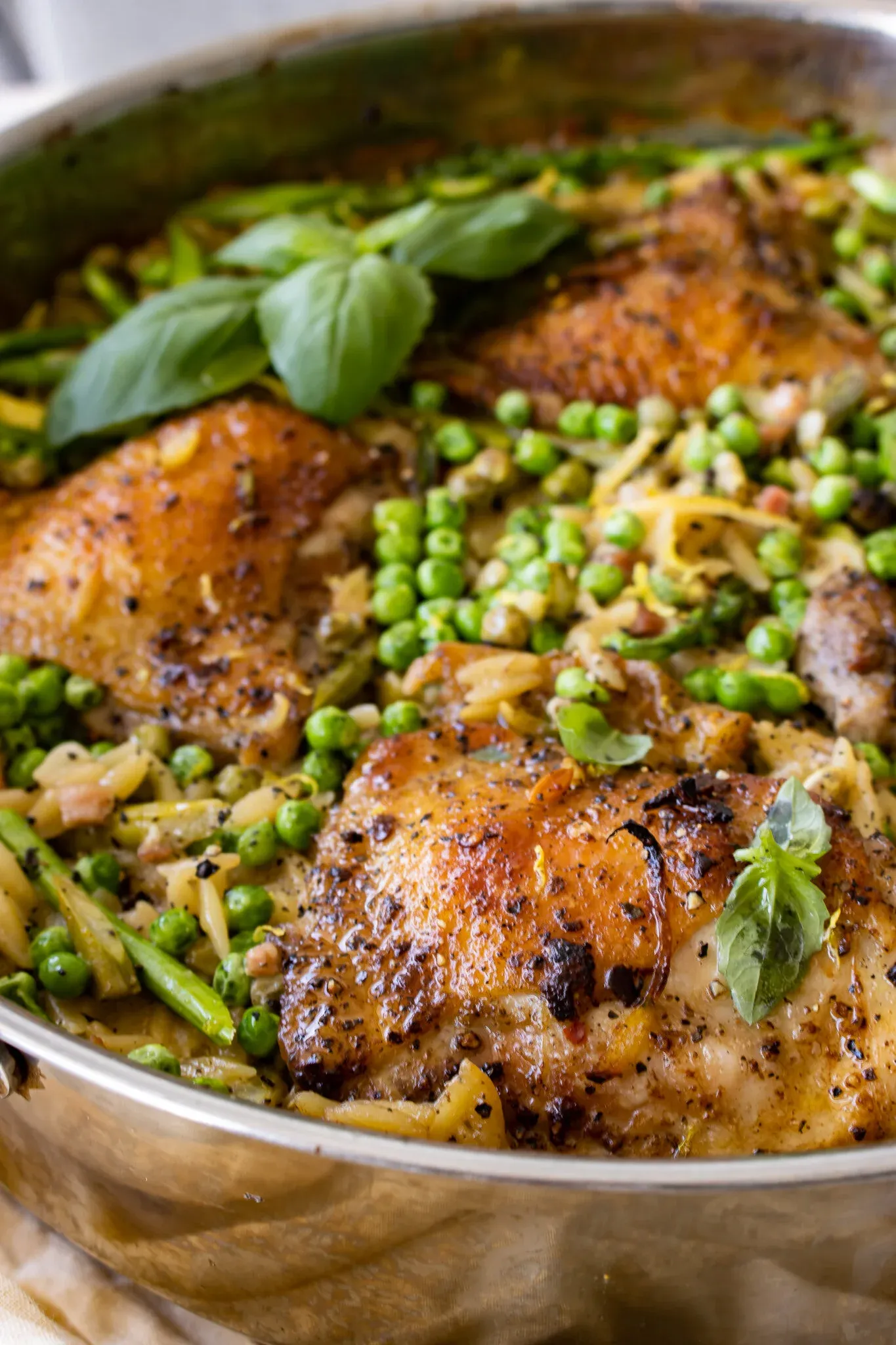 Angled close-up of three pepper-seasoned golden chicken thighs in the stainless skillet surrounded by bright green peas