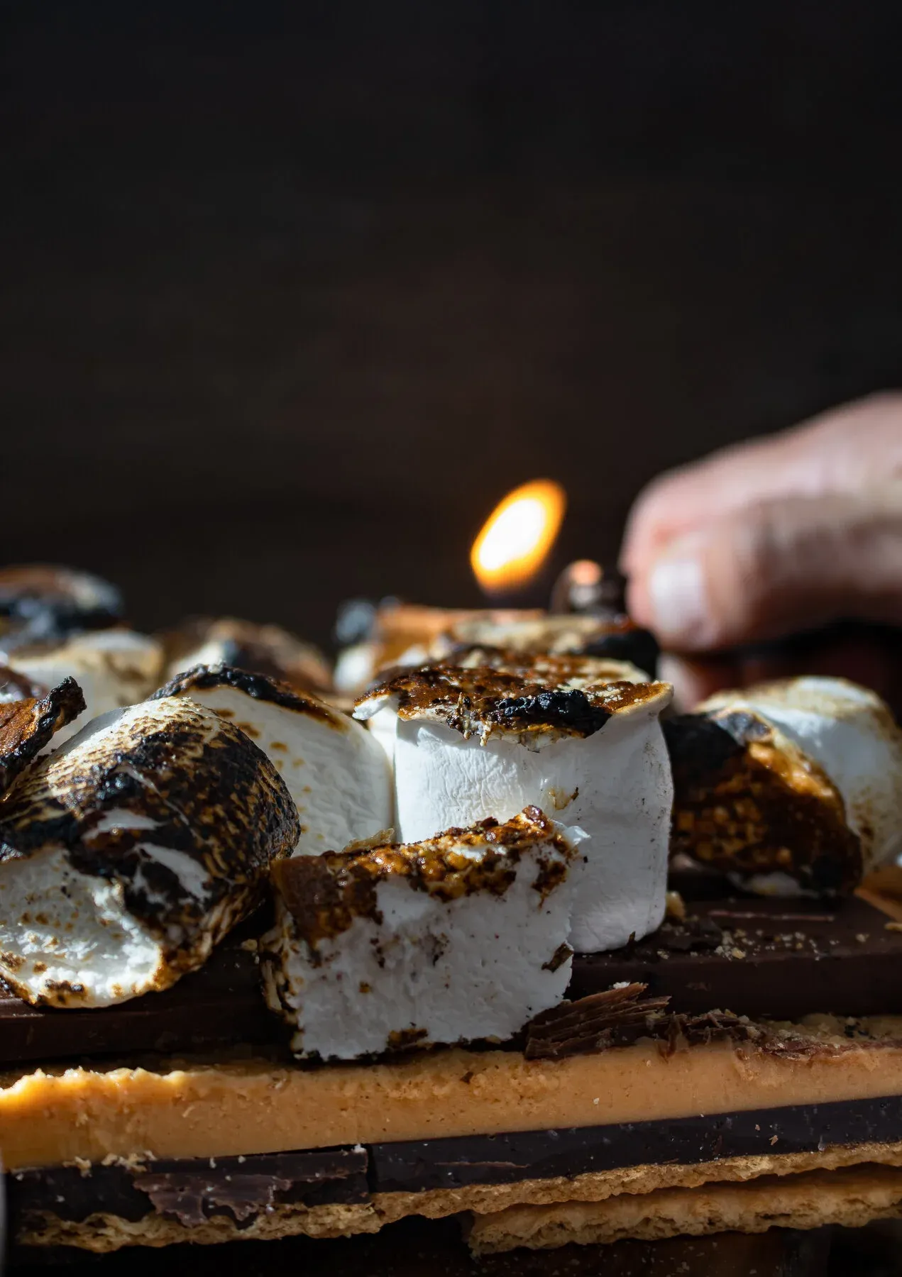 Dramatic dark studio shot of a hand holding a lighter with an open flame above a slab of the no-bake s'mores bars