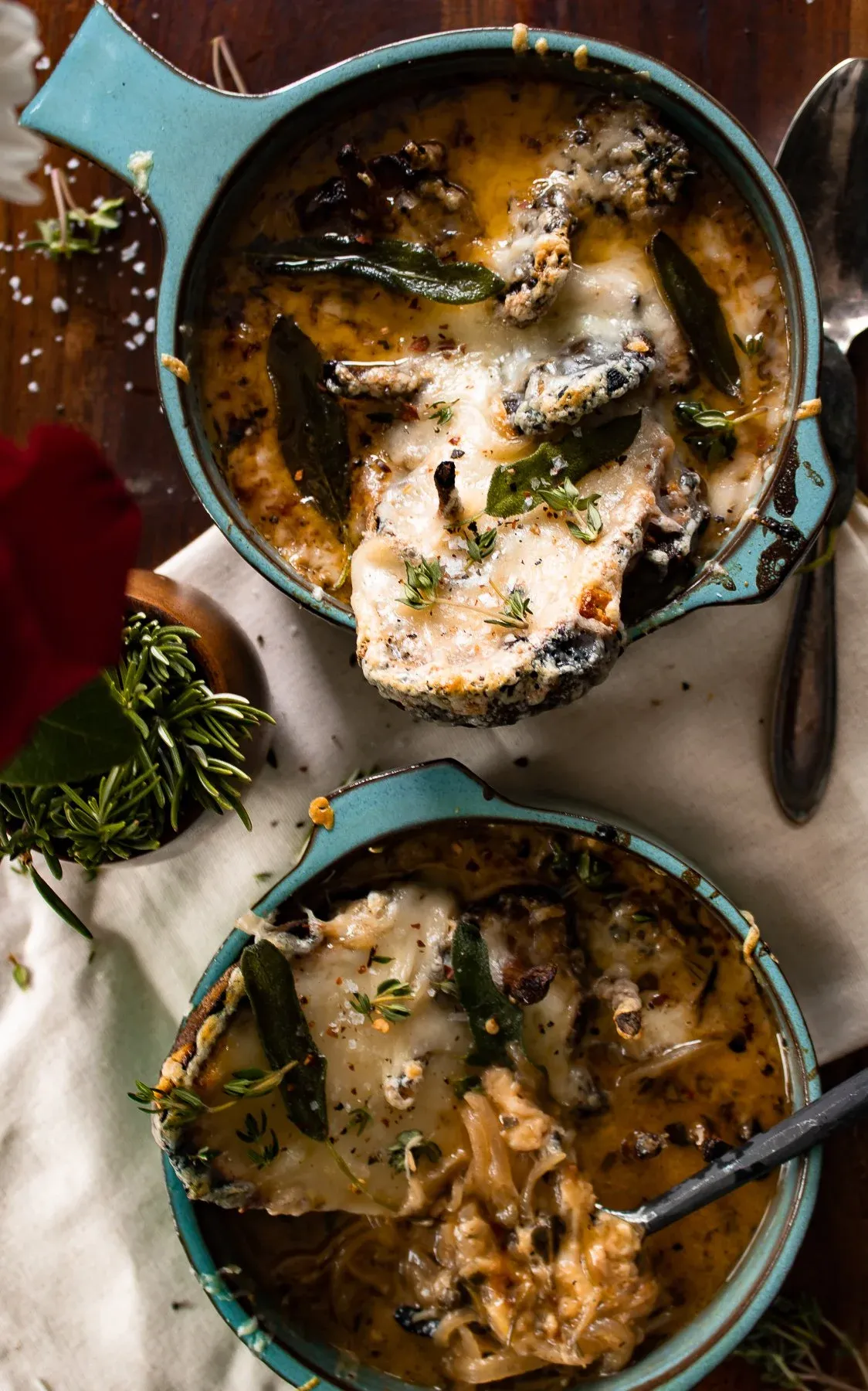 Slightly elevated overhead view of two teal handled ceramic crocks of French Onion Soup with Roasted Mushrooms side by side on a linen cloth and dark wood surface