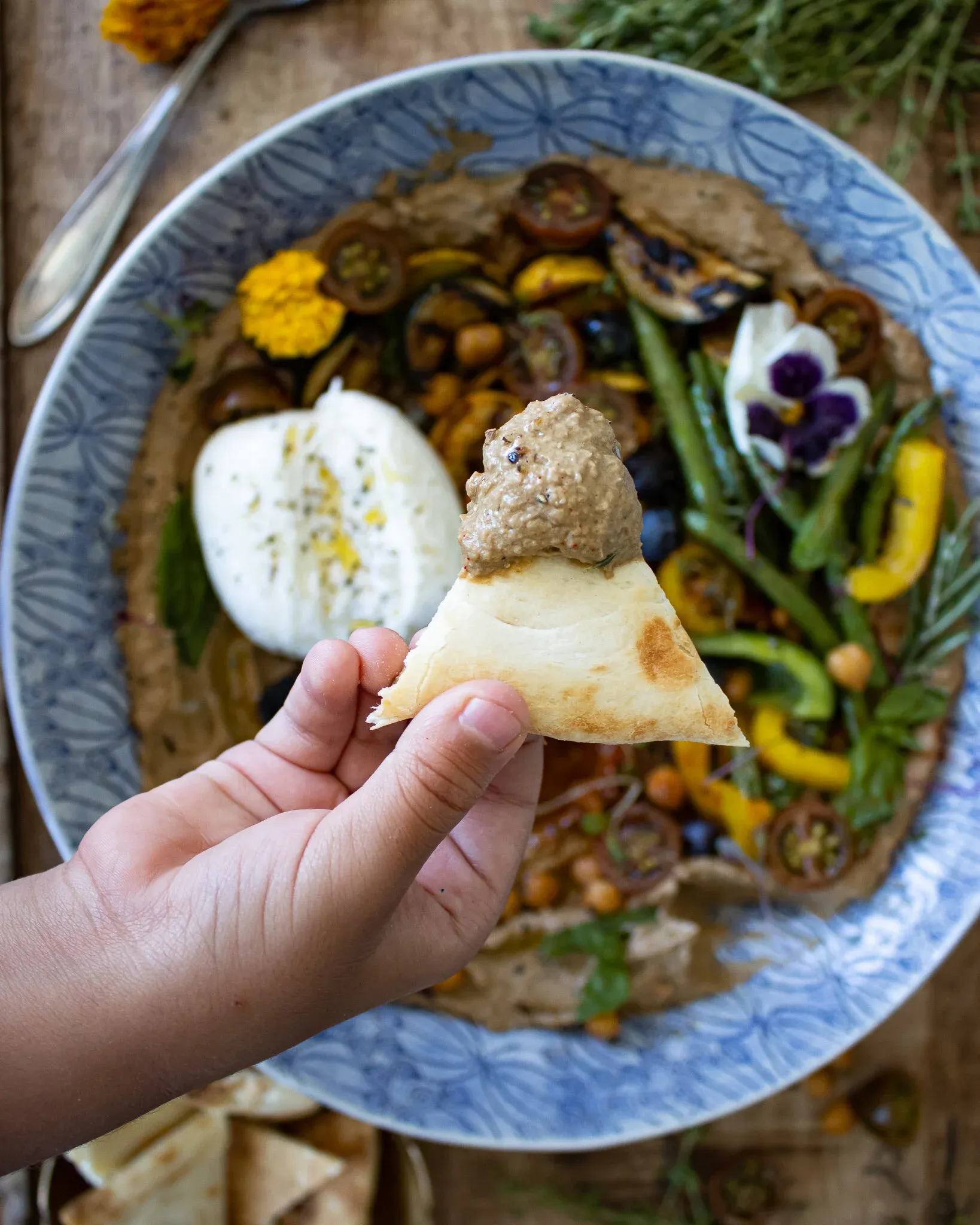 A hand holds a warm pita triangle generously scooped with dark black garlic hummus in the foreground