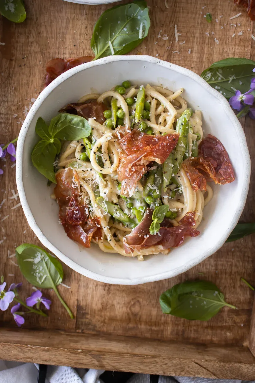 Overhead view of Cacio e Pepe with Spring Veggies and Crispy Prosciutto in a white ceramic bowl on a carved wood tray