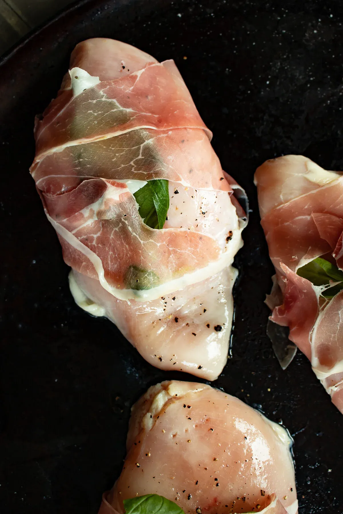 Raw prosciutto-wrapped chicken pieces in a dark cast iron skillet before cooking