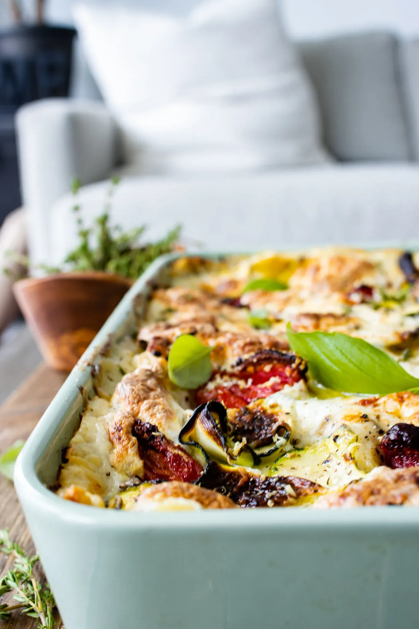 Angled lifestyle shot of the Spinach, Zucchini and Sun-dried Tomato Lasagna in its sage-green ceramic baking dish resting on a light wood board