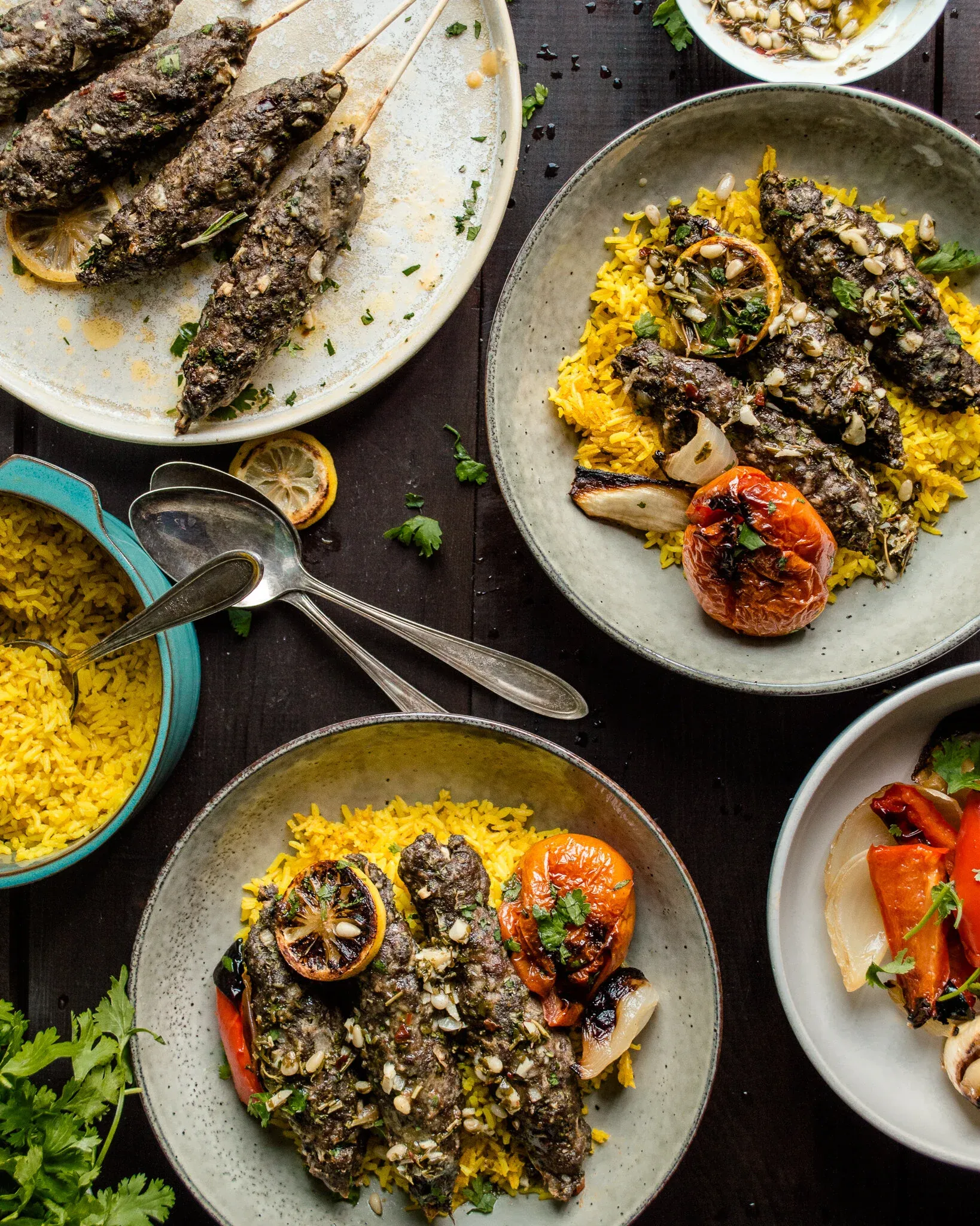 Overhead spread of lamb and beef kabob meal components on a dark wood table: a platter of kabobs at upper left