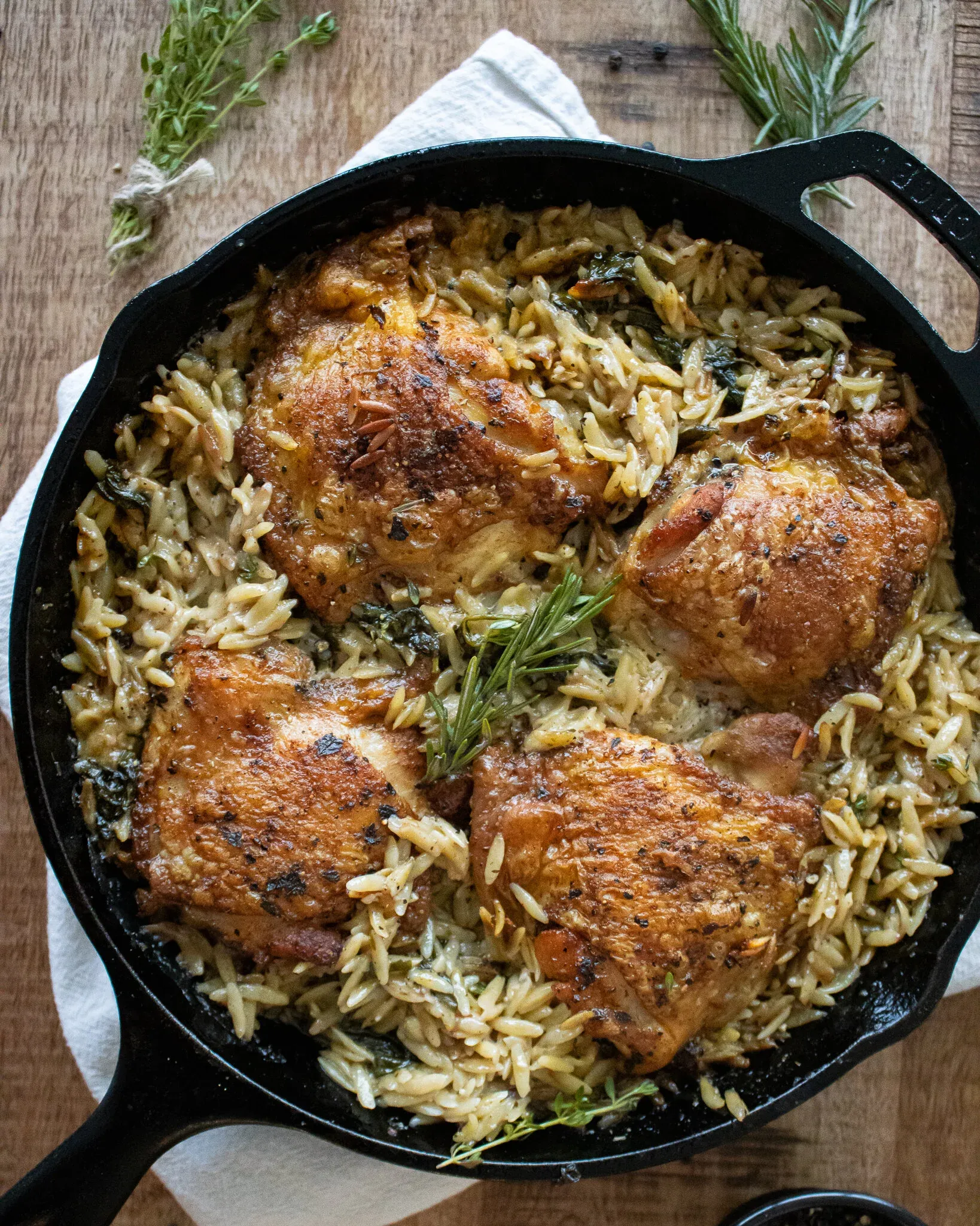 Top-down view of the cast iron skillet holding four golden-skinned chicken thighs on herb-flecked orzo with wilted spinach and a rosemary garnish