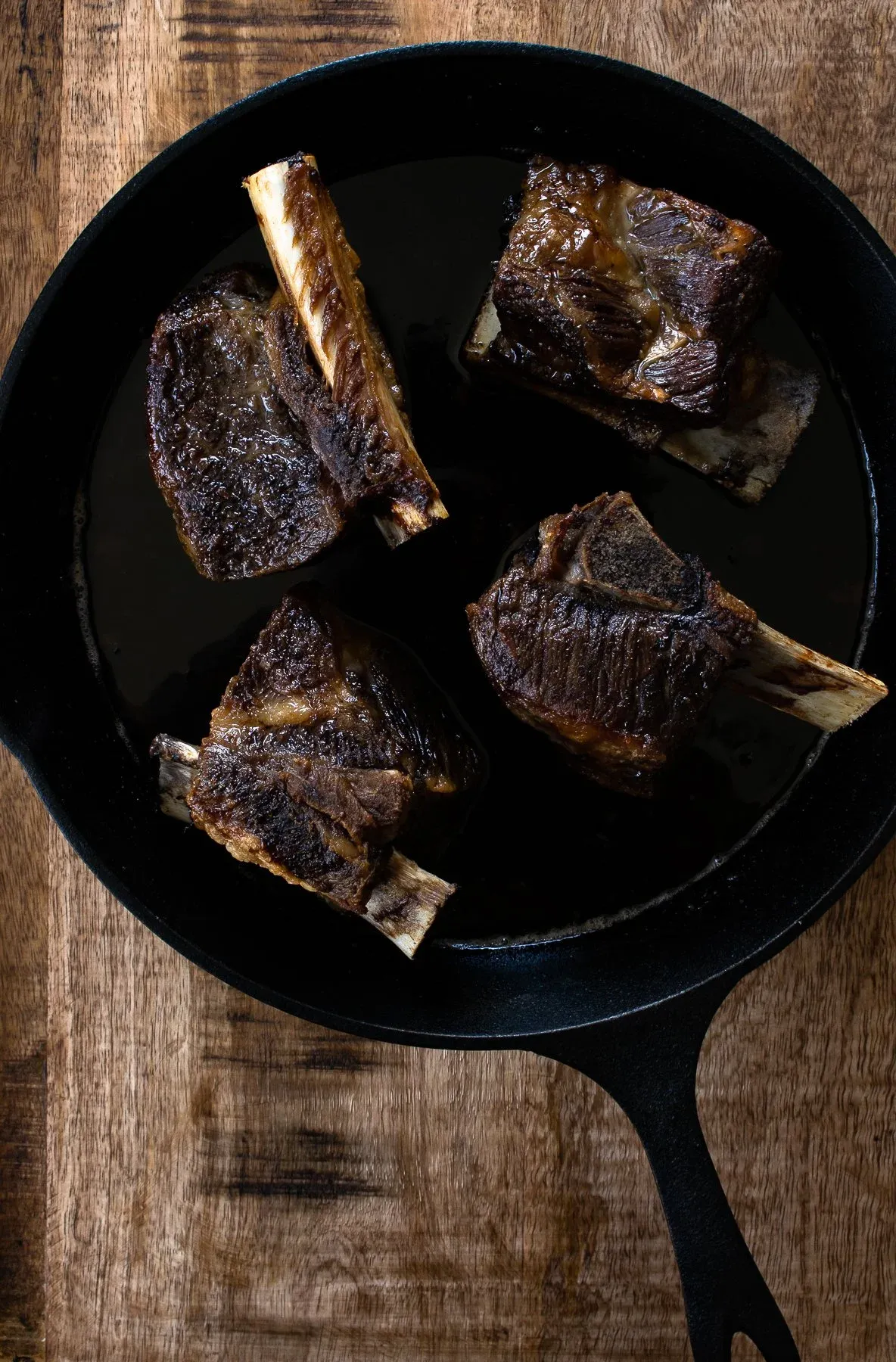 Four deeply seared bone-in beef short ribs with dark mahogany crust arranged in a black cast iron skillet