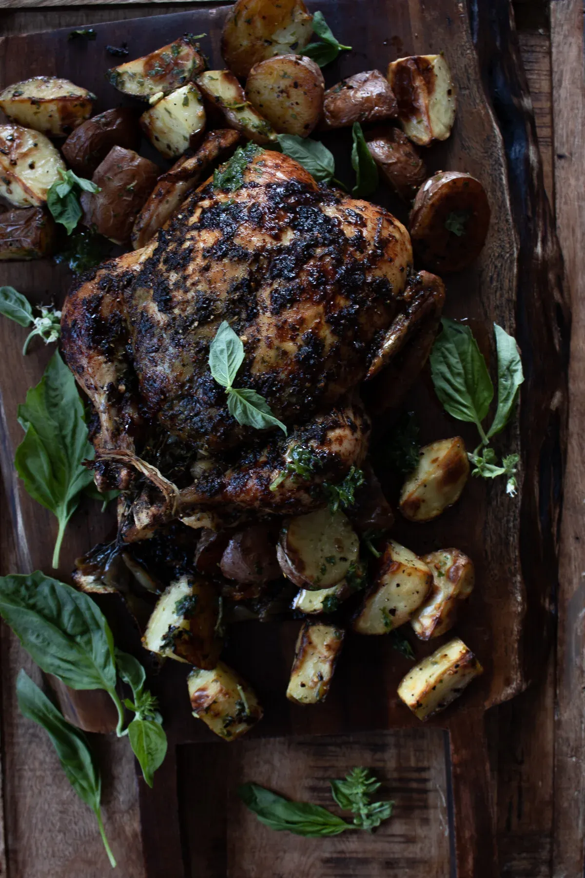 Grilled whole chicken with blackened herb-marinated skin resting on a wide rustic wood cutting board surrounded by chunky roasted herb potatoes and large fresh basil leaves