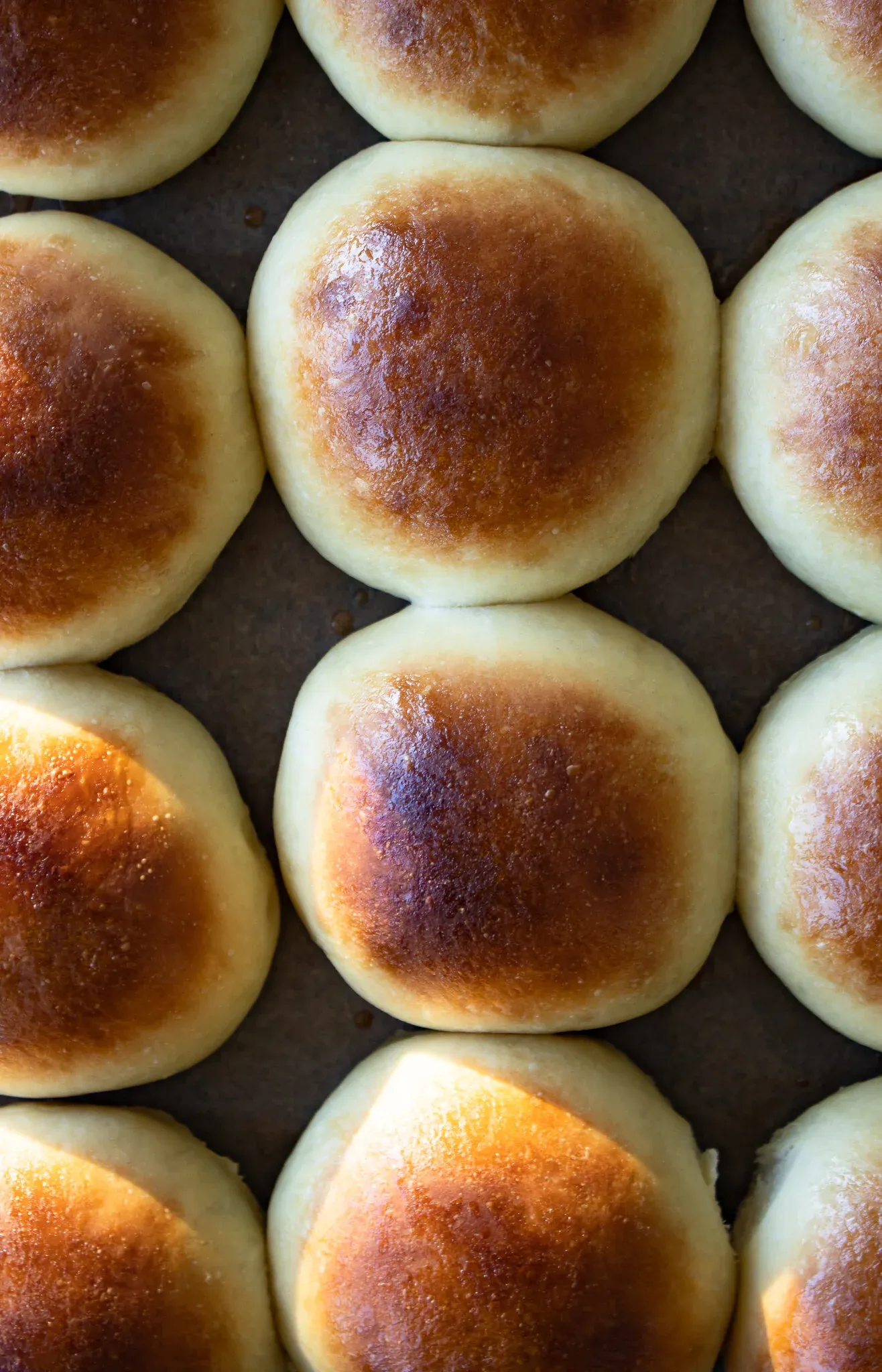 Freshly baked slider buns arranged in tight rows on a dark baking sheet, tops golden-brown with deep caramelized spots