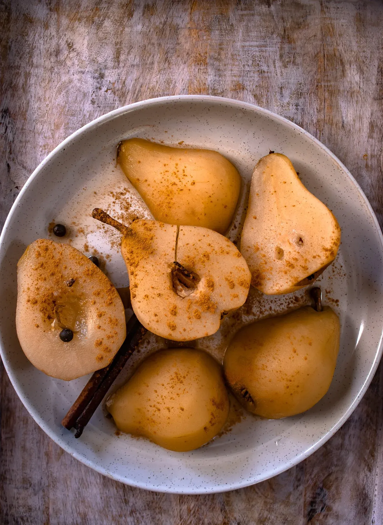 Overhead view of six spiced poached pear halves arranged in a large shallow white speckled ceramic bowl filled with golden amber poaching syrup