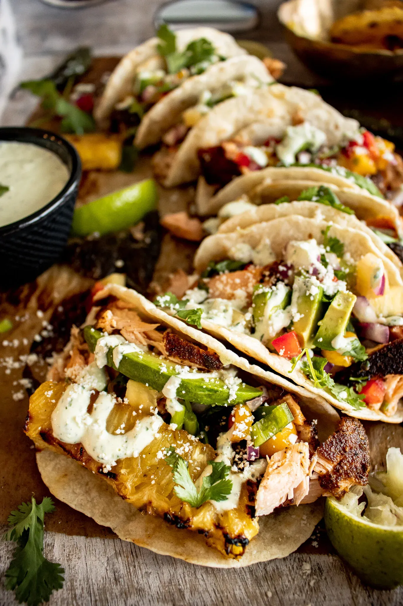 Side-angle close-up of a Blackened Salmon Taco in the foreground overflowing with grilled pineapple chunks, sliced avocado