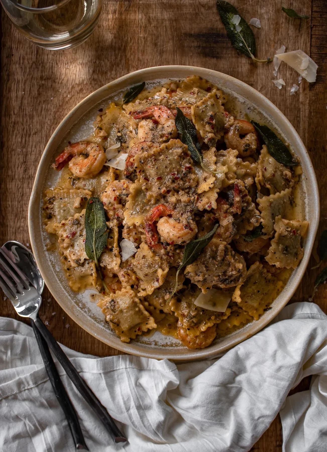 Wider overhead view of the complete plating of Sage and Browned Butter Shrimp Scampi with Ravioli in a cream ceramic bowl on a dark wood table