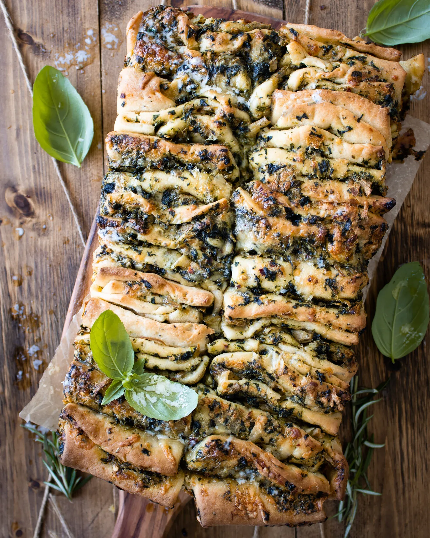 Overhead flat-lay of pesto spinach and artichoke pull-apart bread cut open into individual portions on a wooden board with parchment paper