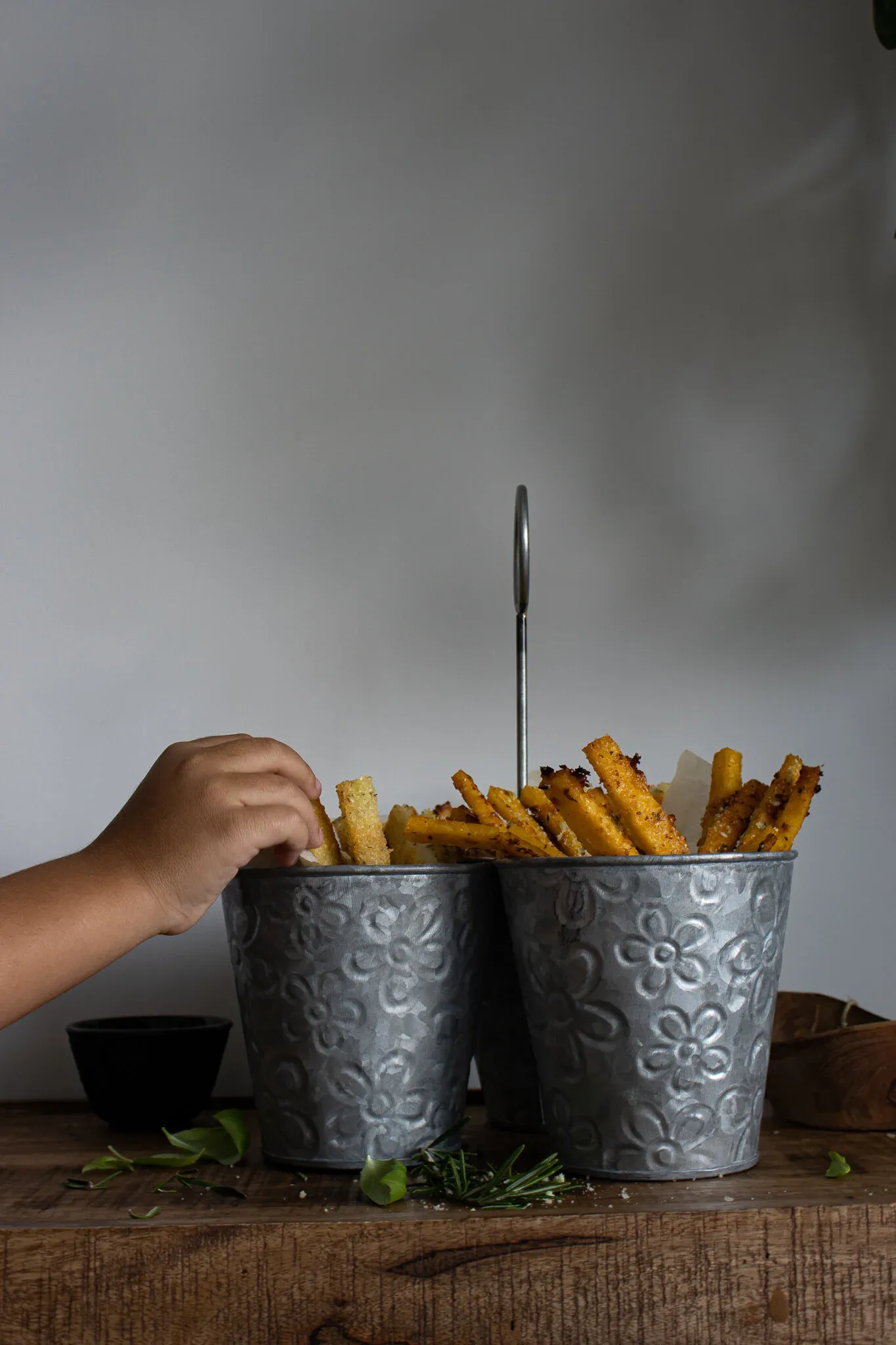 Side-angle view of parmesan herb polenta fries standing upright in two silver embossed floral galvanized metal pails connected by a loop handle on a wooden surface