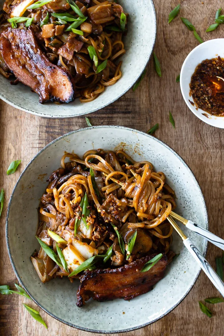 Overhead flat-lay of two plates of chili crisp noodles with pan-fried pork belly