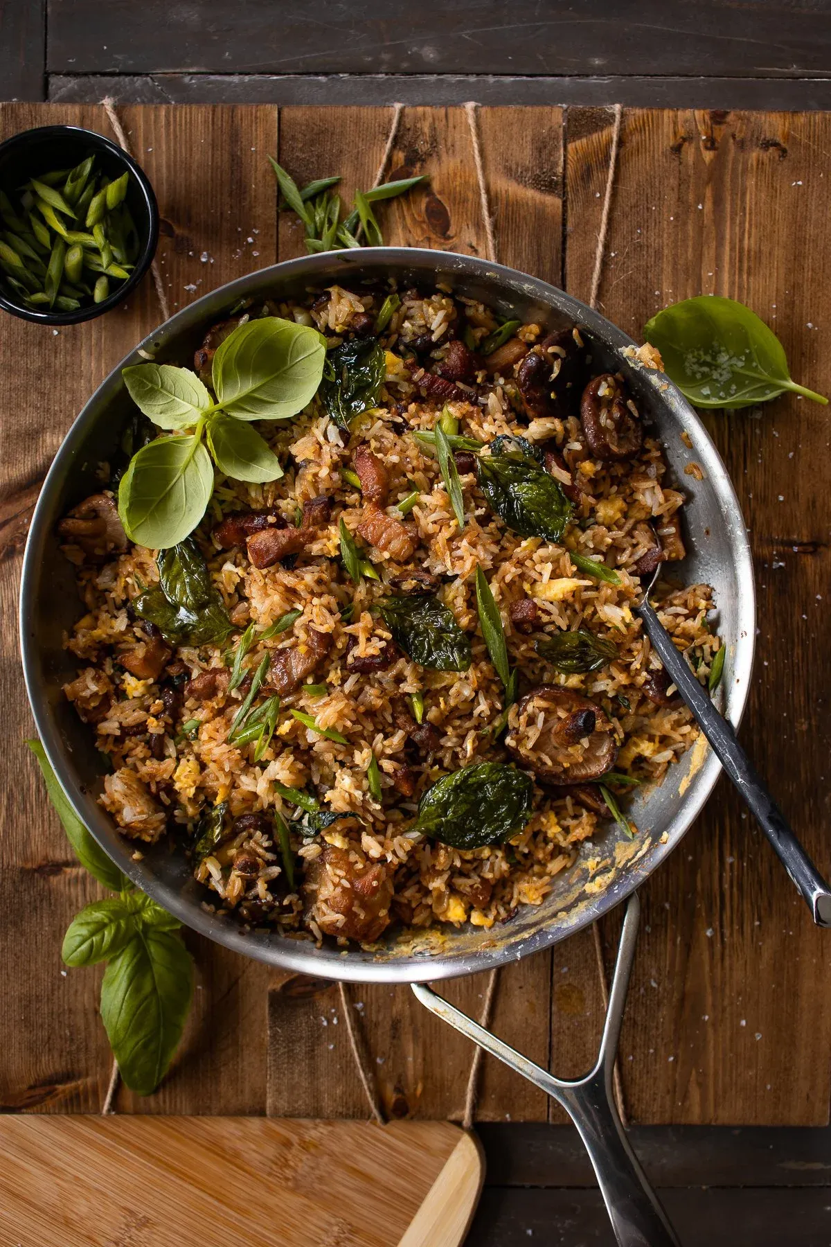 Wider overhead shot of the pork belly, mushroom and crispy basil fried rice in a stainless steel skillet centered on a dark wood cutting board