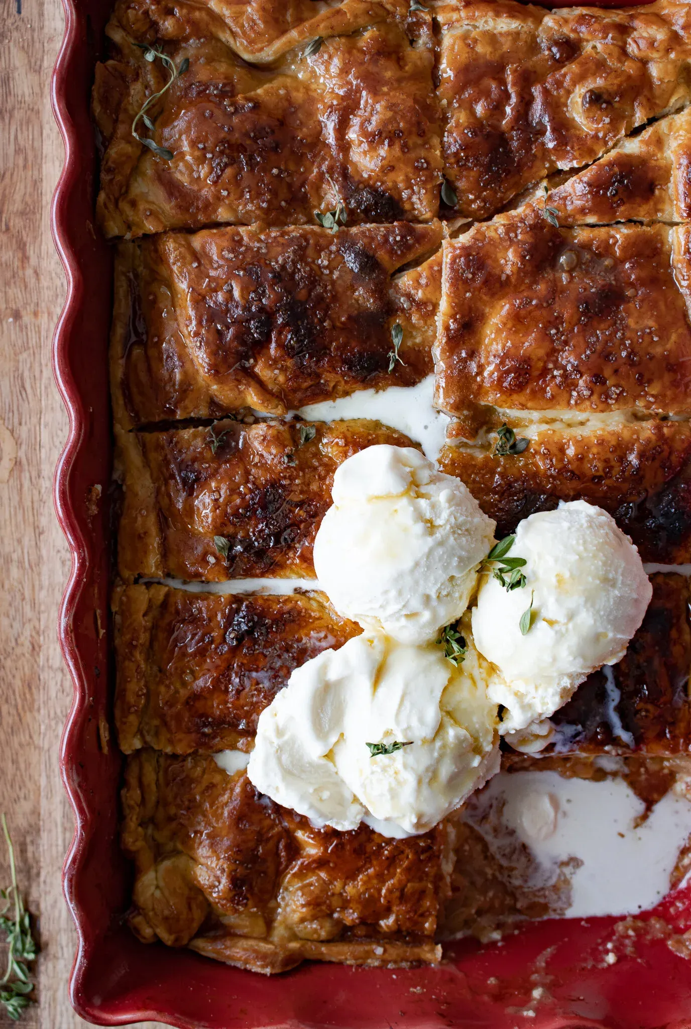 Close-up overhead shot of Apple and Bourbon Pandowdy with three scoops of vanilla ice cream melting over the dark golden caramelized puff pastry squares in a red ceramic baking dish