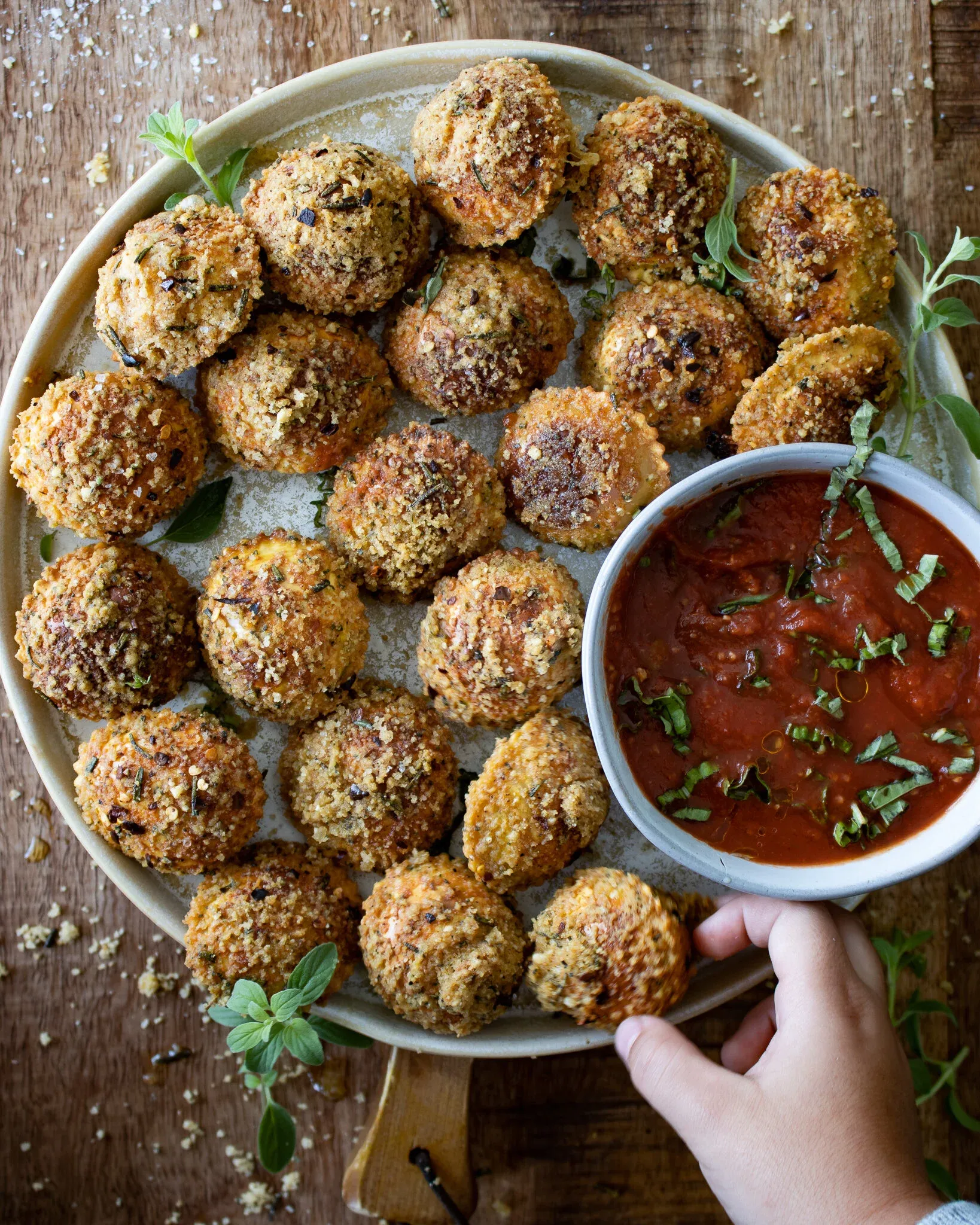 Overhead shot of the oven-baked fried ravioli platter with a hand extending from the lower right corner holding the small white bowl of bright red marinara sauce flecked with fresh chiffonade basil