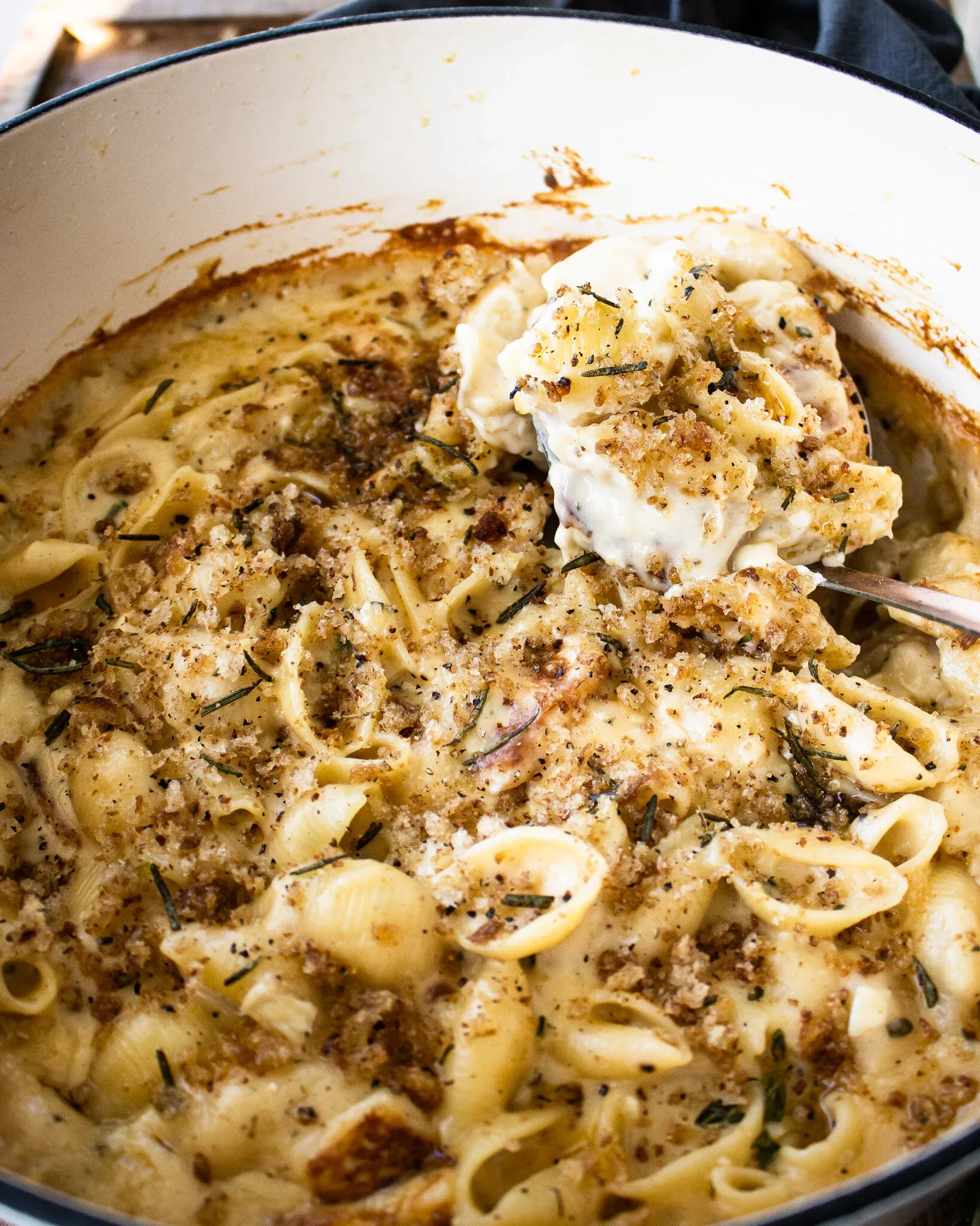 Close-up angled shot of a large spoon lifting a serving of 7 Cheese Herb and Garlic Baked Mac and Cheese from the white enamel pot