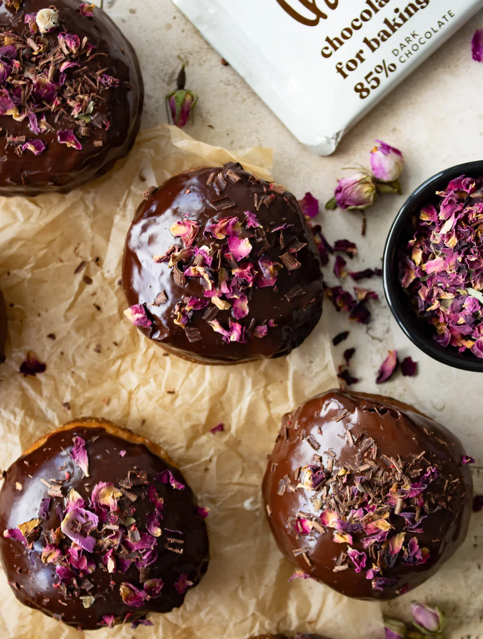 Overhead close-up of four cardamom and rose brioche doughnuts on crinkled parchment with their glossy dark chocolate glaze catching the light