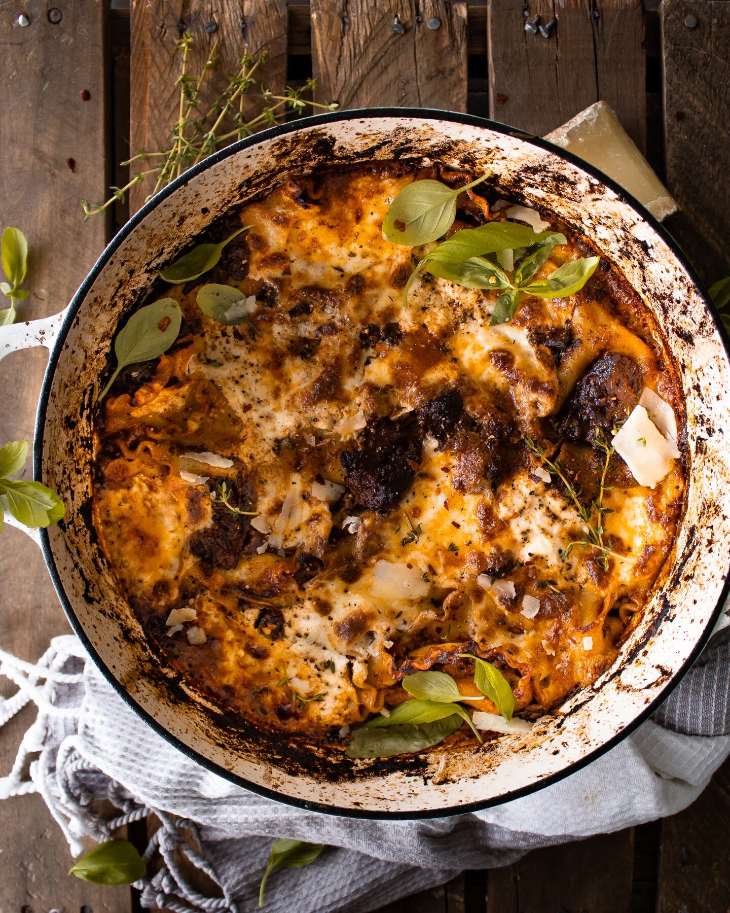 Overhead view of Short Rib Skillet Lasagna in a round white enamel braiser with handles, on a dark wooden pallet surface