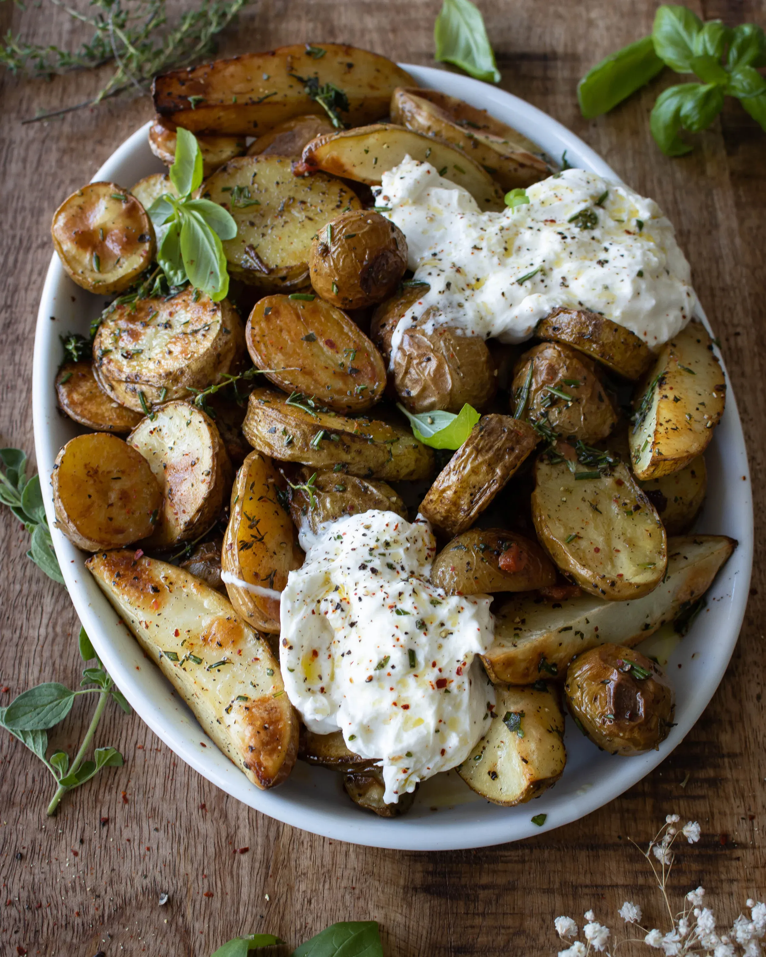 Wide overhead shot of the finished Roasted Herby Potatoes with Burrata in a white round bowl on a wooden board