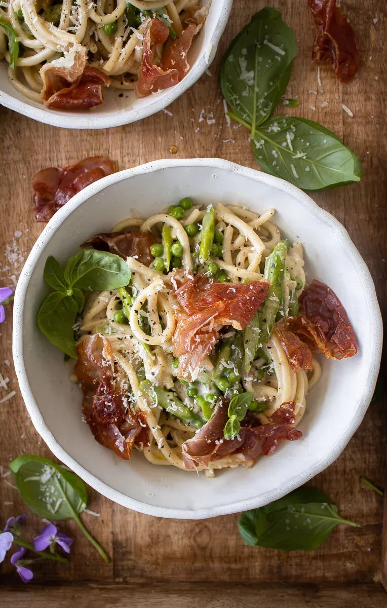 Overhead view of two white bowls of Cacio e Pepe with Spring Veggies and Crispy Prosciutto on a wood surface
