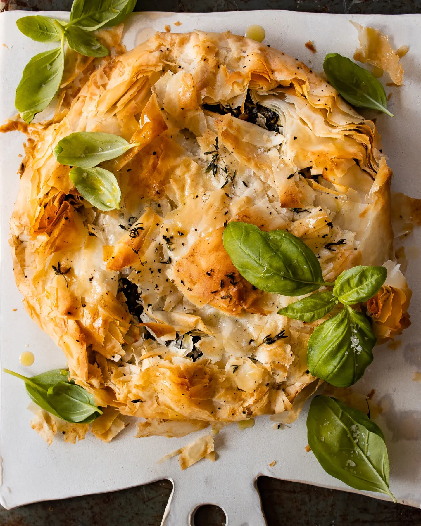 Overhead view of the whole round Spinach and Feta Phyllo Pie on a white ceramic paddle board against a dark surface