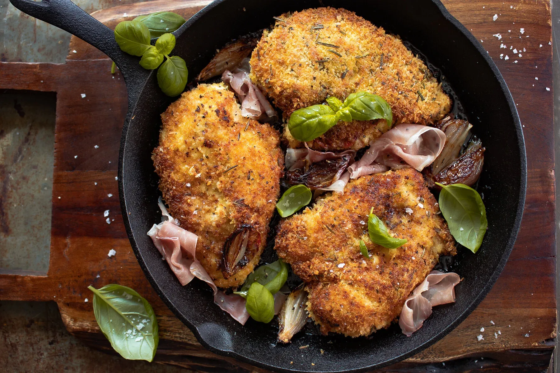 Wide overhead shot of all three Prosciutto and Basil Wrapped Chicken cutlets in the black cast iron skillet on a dark wooden board