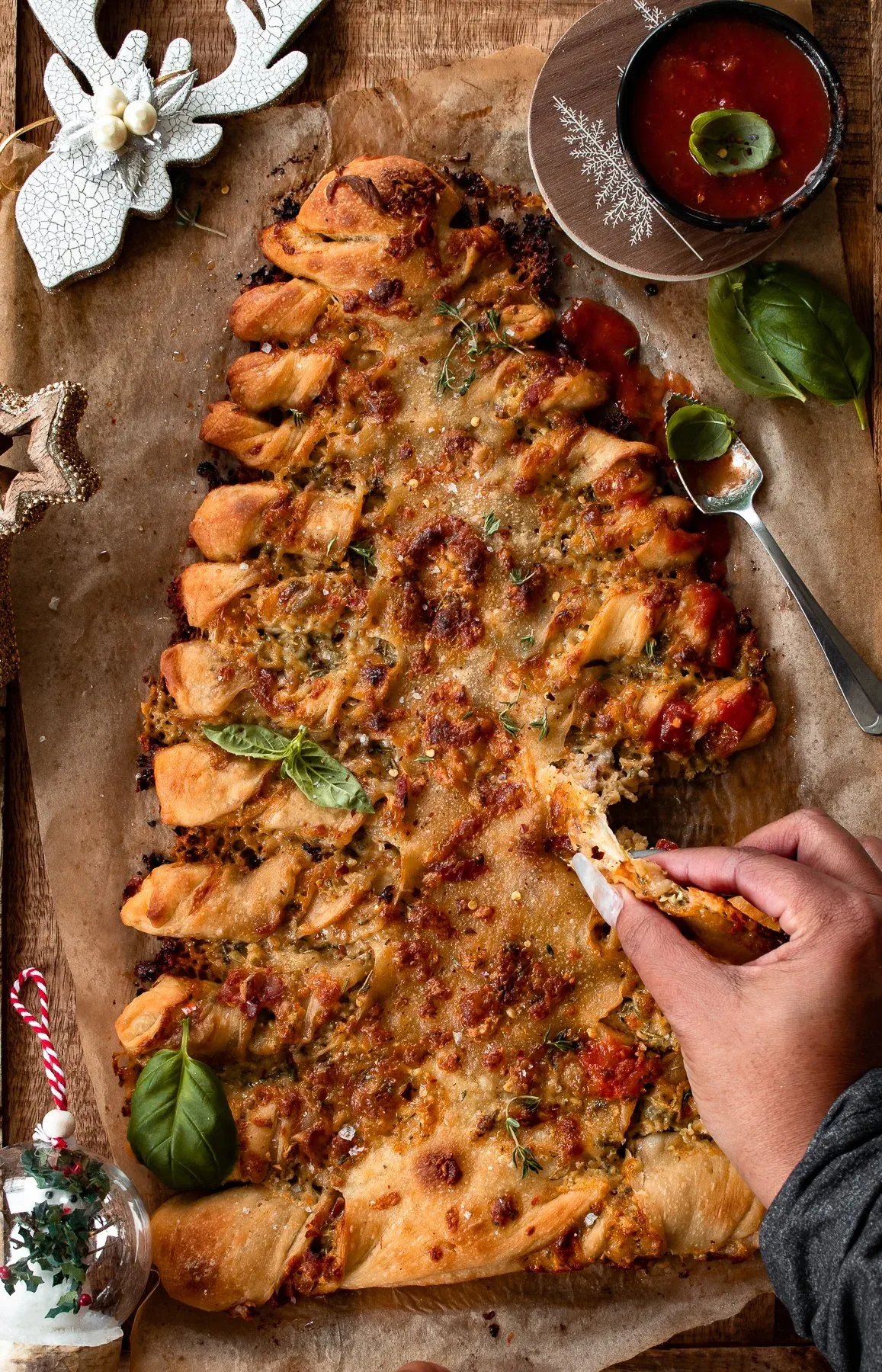 A dark-sleeved hand reaching in from the right to pull off a piece of the Christmas Tree Pull Apart Bread from a baking sheet lined with parchment
