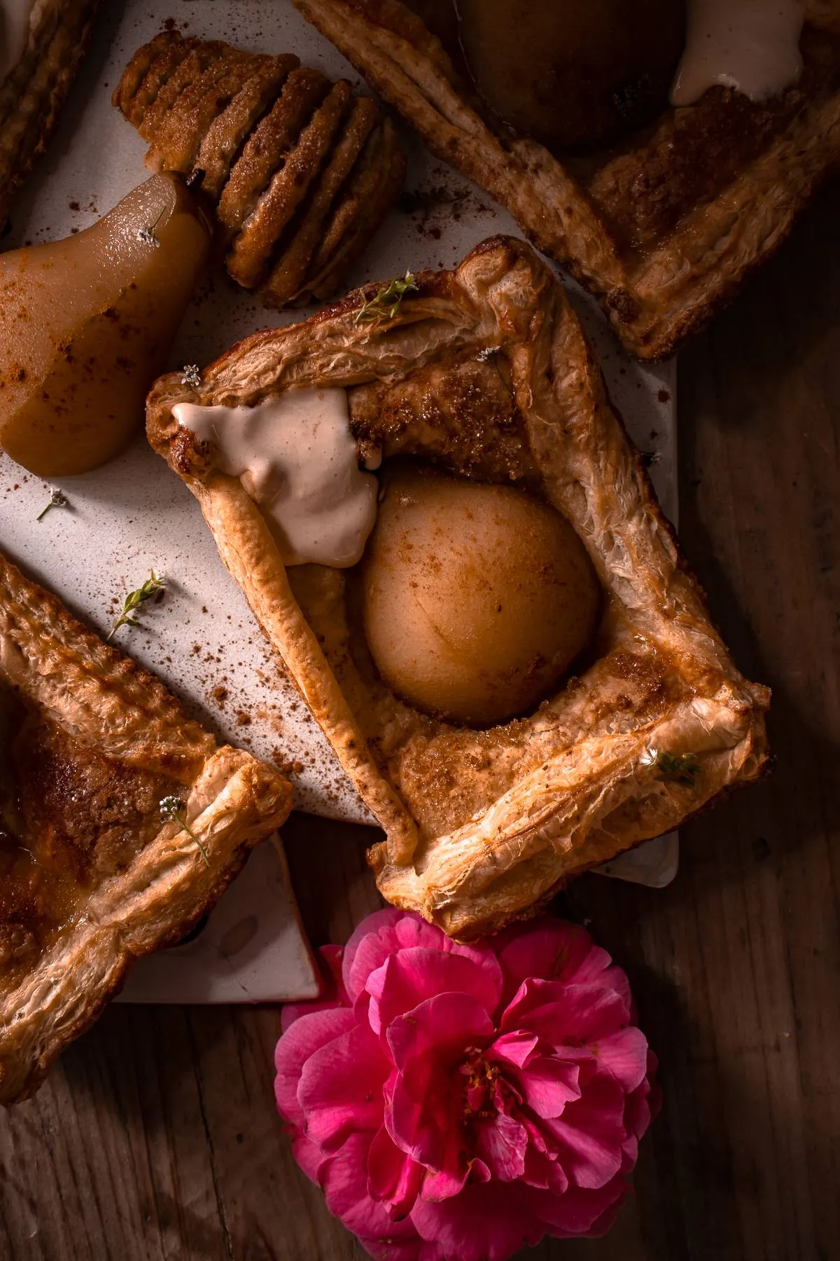 Overhead dark-moody flat-lay of five rectangular poached pear pastry tarts with mascarpone cream arranged on a white ceramic surface atop a dark wood table