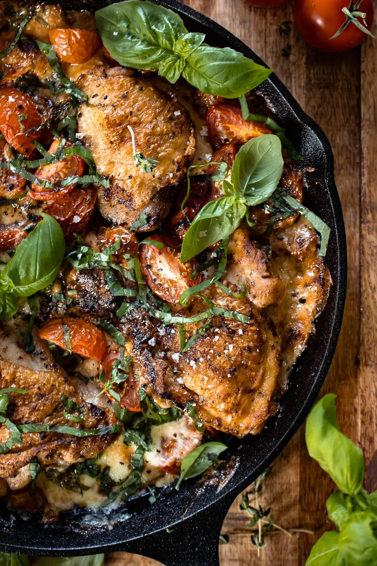 Overhead view of cast iron caprese chicken thighs in a black cast iron skillet on a rustic wooden board