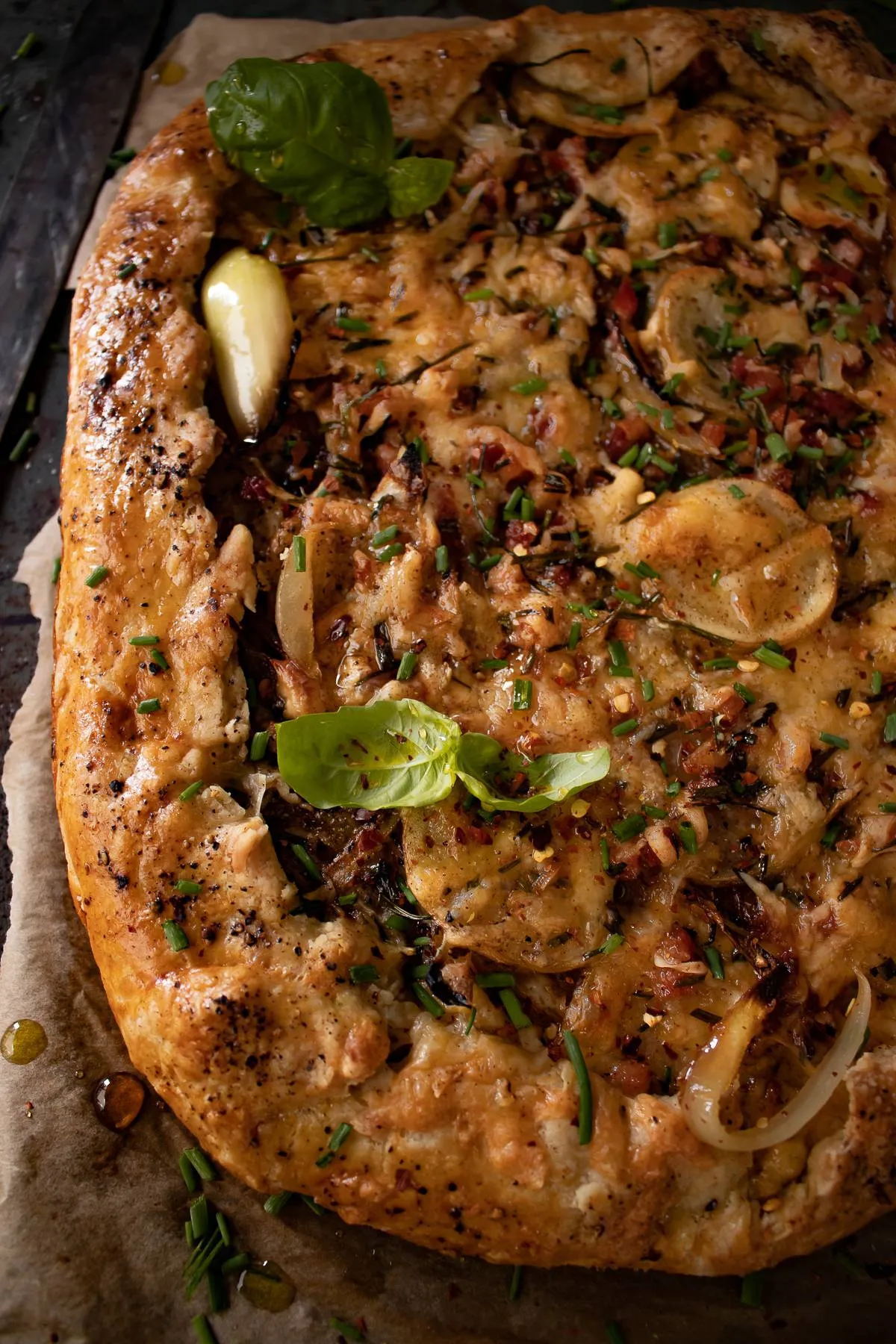 Overhead view of a whole cheesy potato, onion and chive tart with pancetta on parchment paper on a dark metal baking sheet