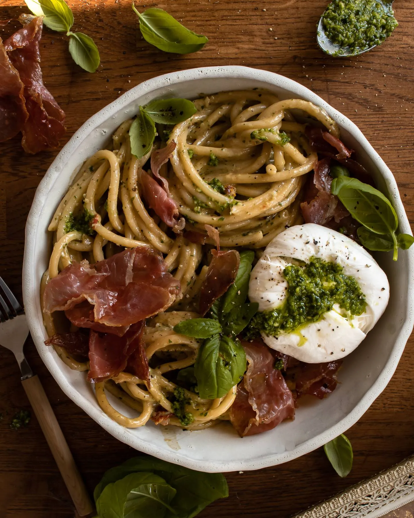Overhead shot of a wide shallow white speckled ceramic bowl on dark wood filled with Creamy Basil and Garlic Pasta with Prosciutto and Burrata
