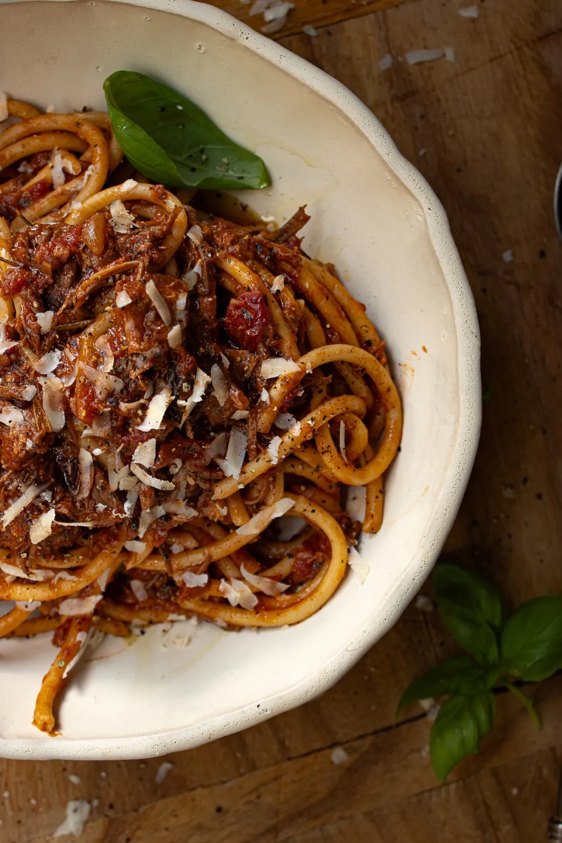 White rustic bowl of Dominican Style Goat Ragù served over thick bucatini pasta coated in a deep-red tomato-braised goat sauce
