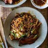 Overhead shot of a speckled off-white ceramic plate of chili crisp noodles with pan-fried pork belly on a light wood surface