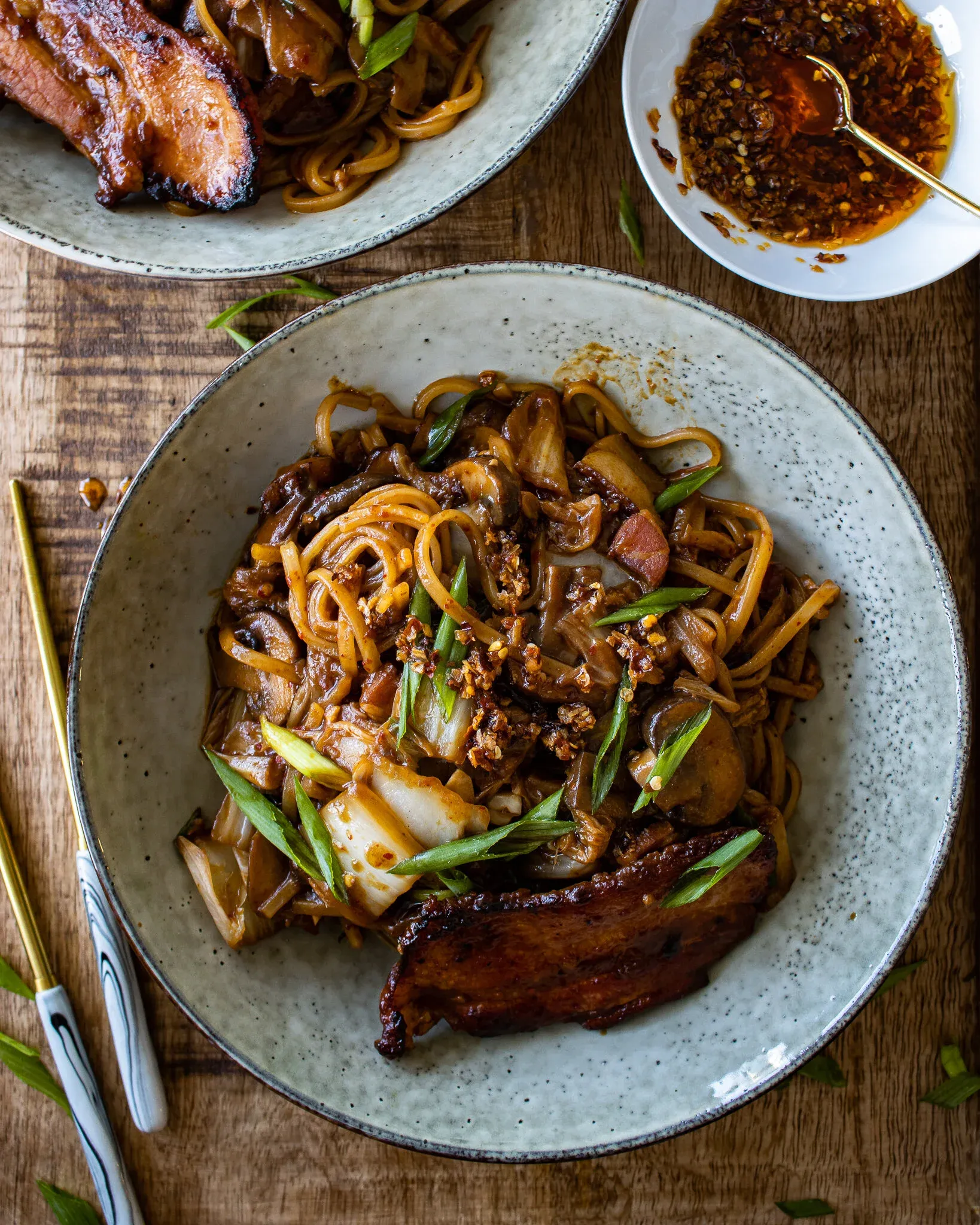Overhead shot of a speckled off-white ceramic plate of chili crisp noodles with pan-fried pork belly on a light wood surface