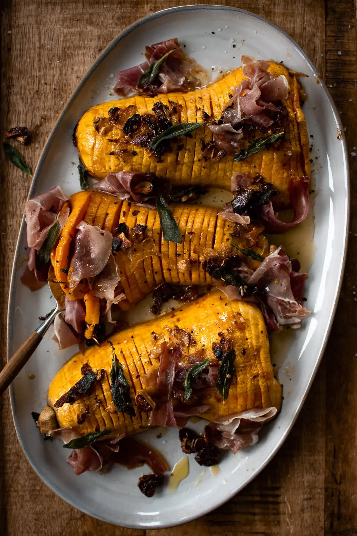 Three caramelized hasselback butternut squash halves arranged on a white oval ceramic platter on a weathered wooden board