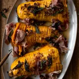 Three caramelized hasselback butternut squash halves arranged on a white oval ceramic platter on a weathered wooden board