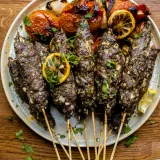Six lamb and beef kabobs on wooden skewers arranged across a round speckled ceramic platter