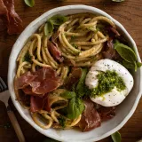 Overhead shot of a wide shallow white speckled ceramic bowl on dark wood filled with Creamy Basil and Garlic Pasta with Prosciutto and Burrata