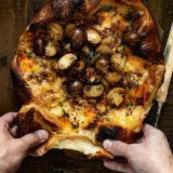 Overhead view of puff pastry wrapped brie with pancetta and mushrooms being torn open by two hands on parchment paper
