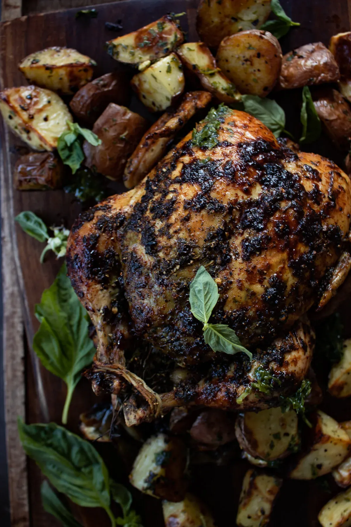 Whole grilled chicken with deeply charred, herb-blackened skin resting on a dark rustic wooden board