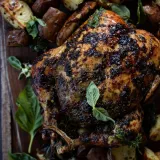 Whole grilled chicken with deeply charred, herb-blackened skin resting on a dark rustic wooden board